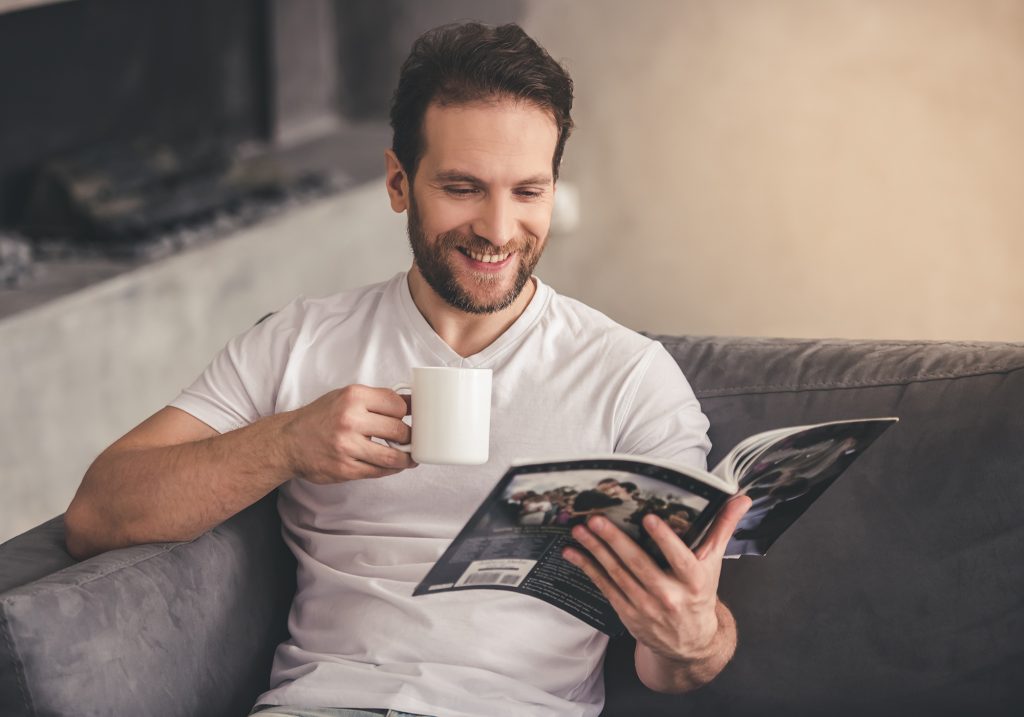 Smiling man sitting on a couch, wearing a white t-shirt, holding a white mug and reading a magazine.